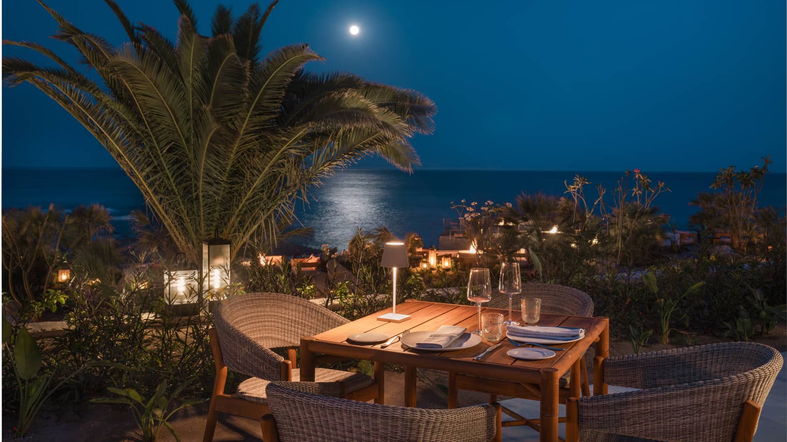 Dining table set for four on an outdoor patio at night with palm trees and the ocean in the distance