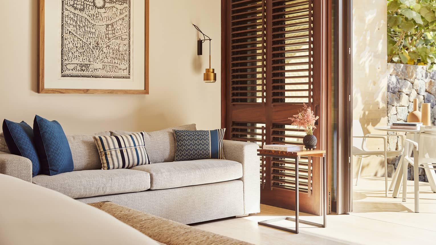 A cozy living space with a light grey sofa adorned with blue and striped pillows. Wooden shutters open to a sunlit patio, and a framed artwork hangs above the sofa.