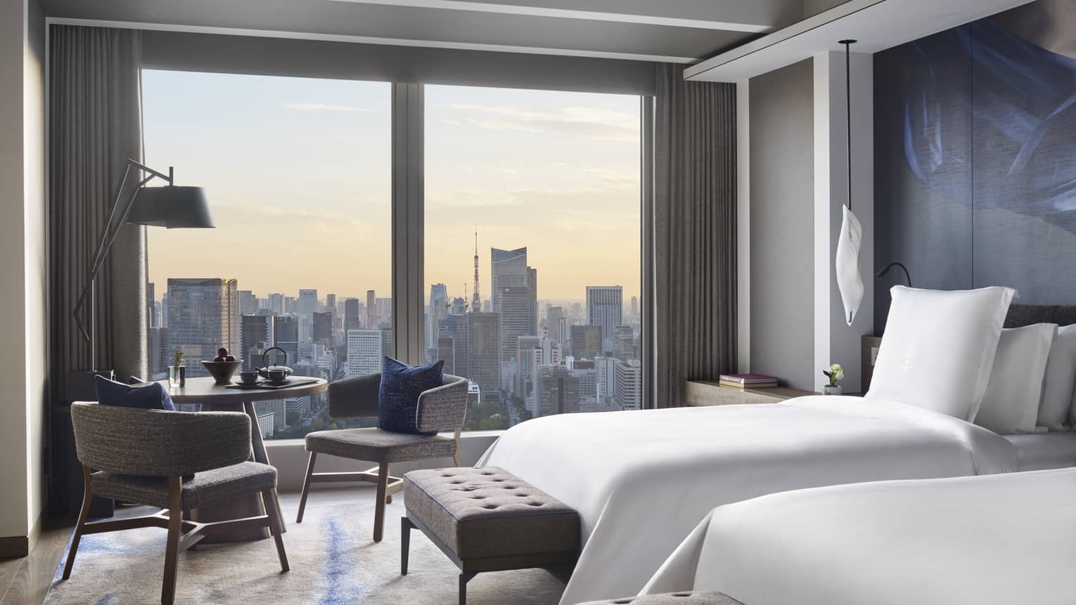 Guest room with two beds, tufted bench, seating area and floor-to-ceiling windows offering panoramic city views