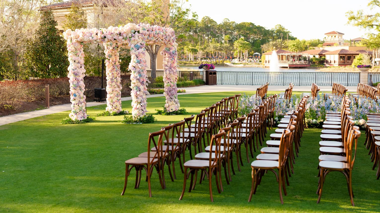 Outdoor wedding set up on a lush green lawn with rows of brown wooden chairs and a large pergola compeltely covered with flowers