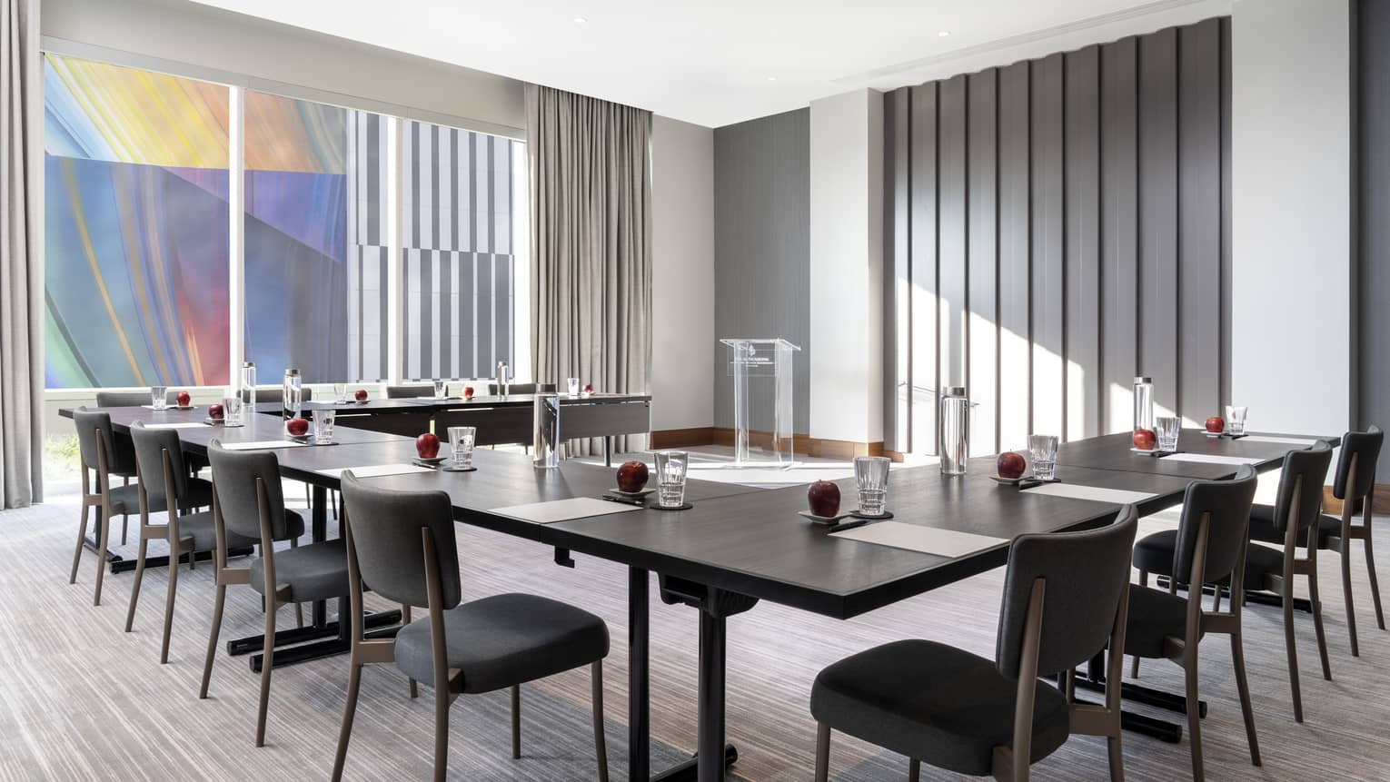 A brightly lit meeting room with a large u-shaped table, dark chairs and a large window.