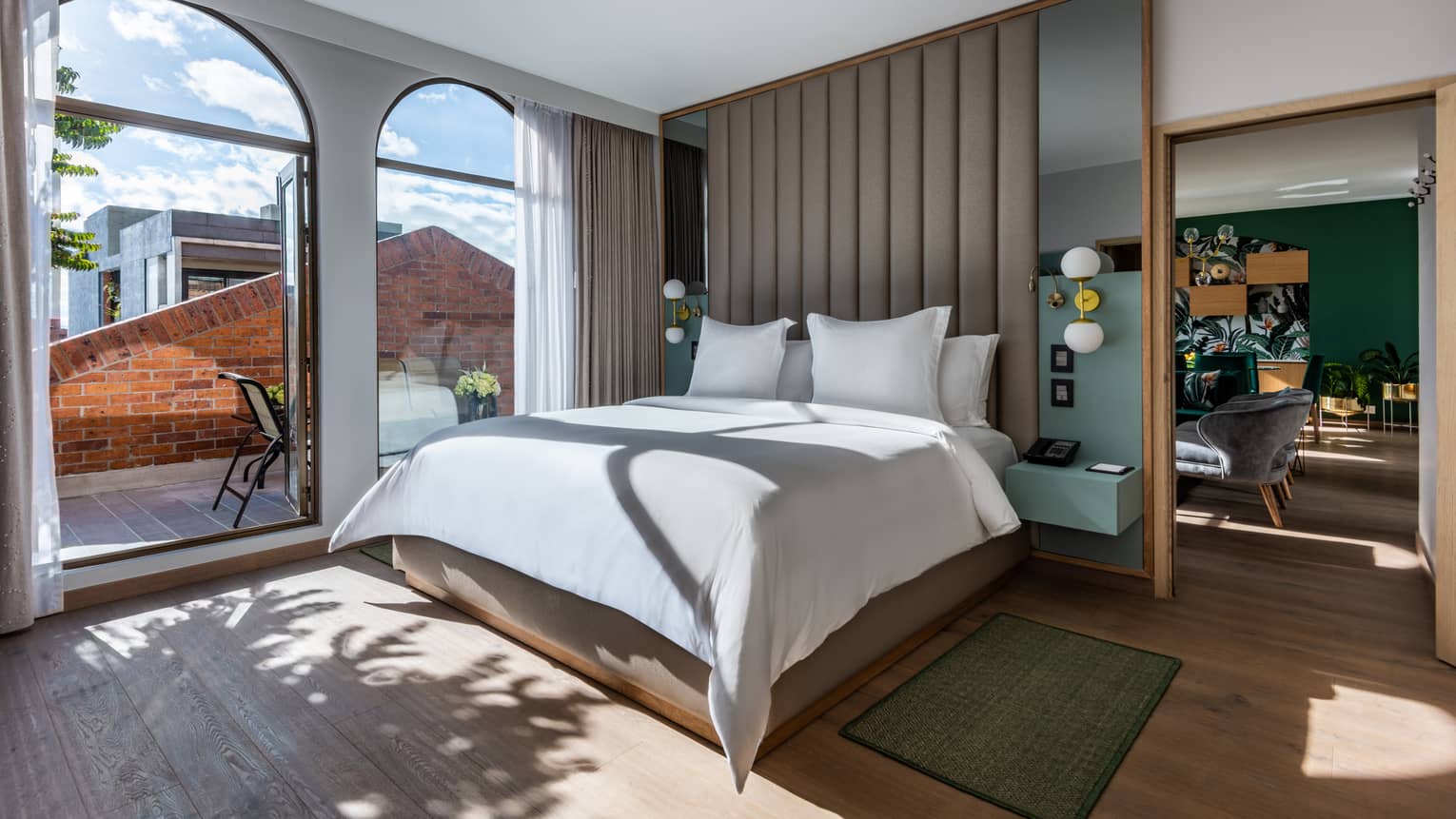 Guest room with bed and white linens, floor-to-ceiling windows overlooking the terrace, living/dining area just visible around the partition
