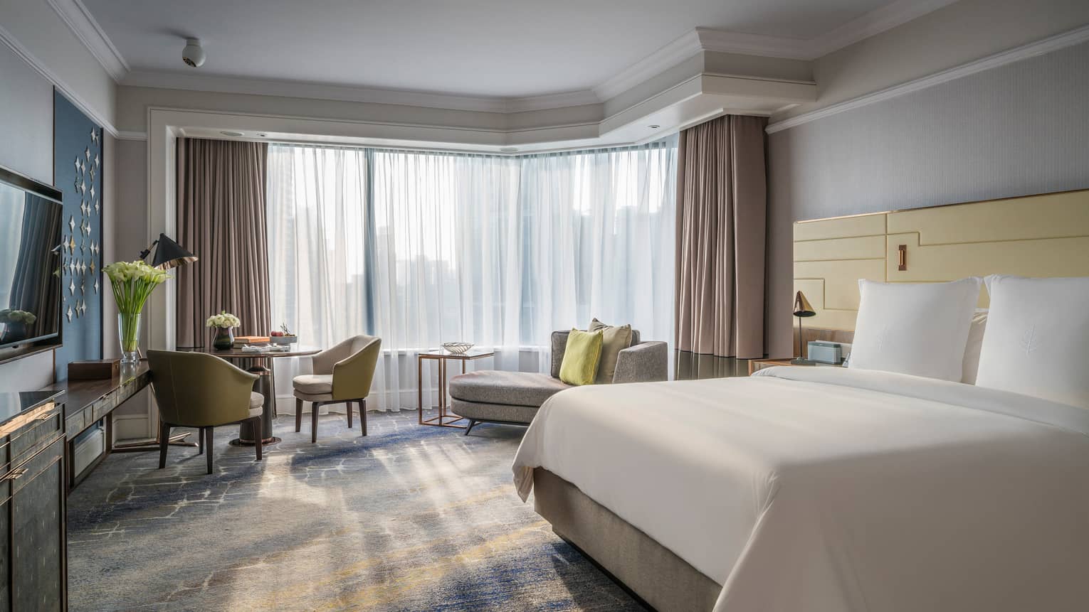 Sunny Deluxe Room bed with white linens, small dining table and chaise lounge, white curtains