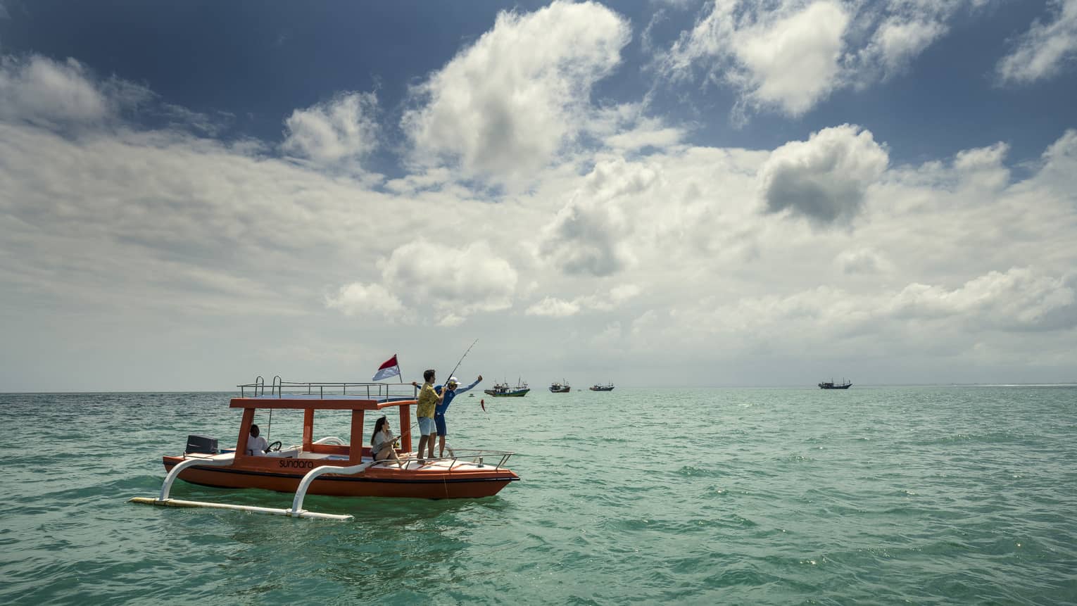 On the open water, three people fish from the hull of an outrigger-style boat; four other boats float in the distance.