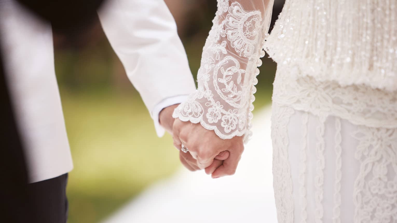 Close-up of bride with lace sleeve and groom with white suit holding hands 