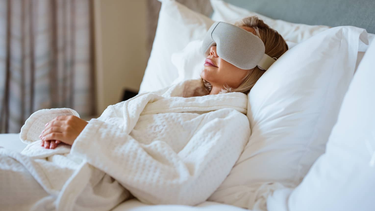 Person wearing white spa robe lays back on a bed with large white pillows wearing a high-tech eye mask