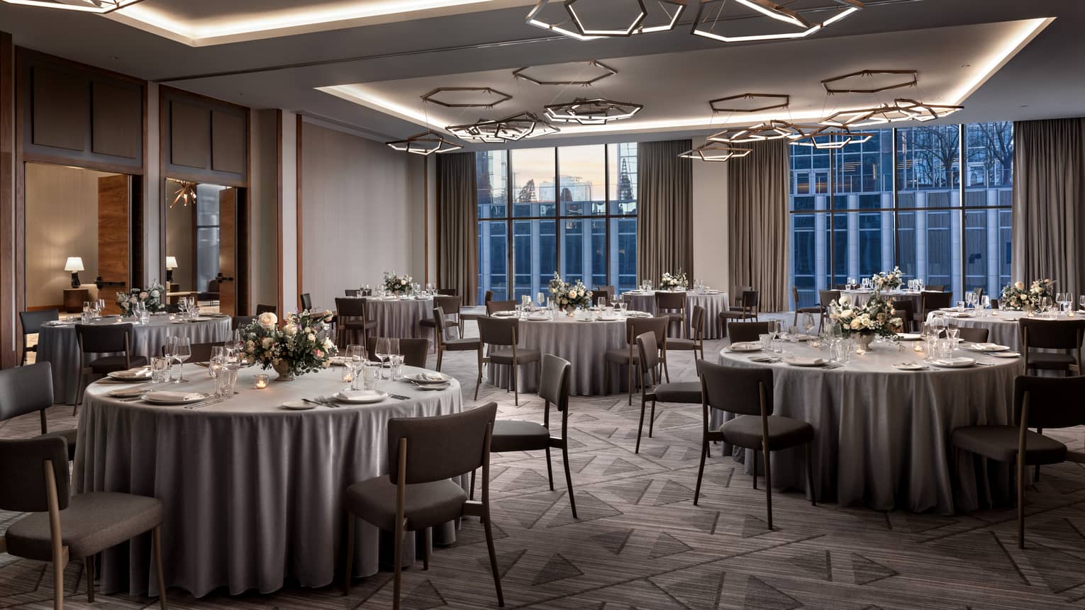 A dimly lit room with round tables, with grey tablecloths and glassware, grey carpet and geometric lights.