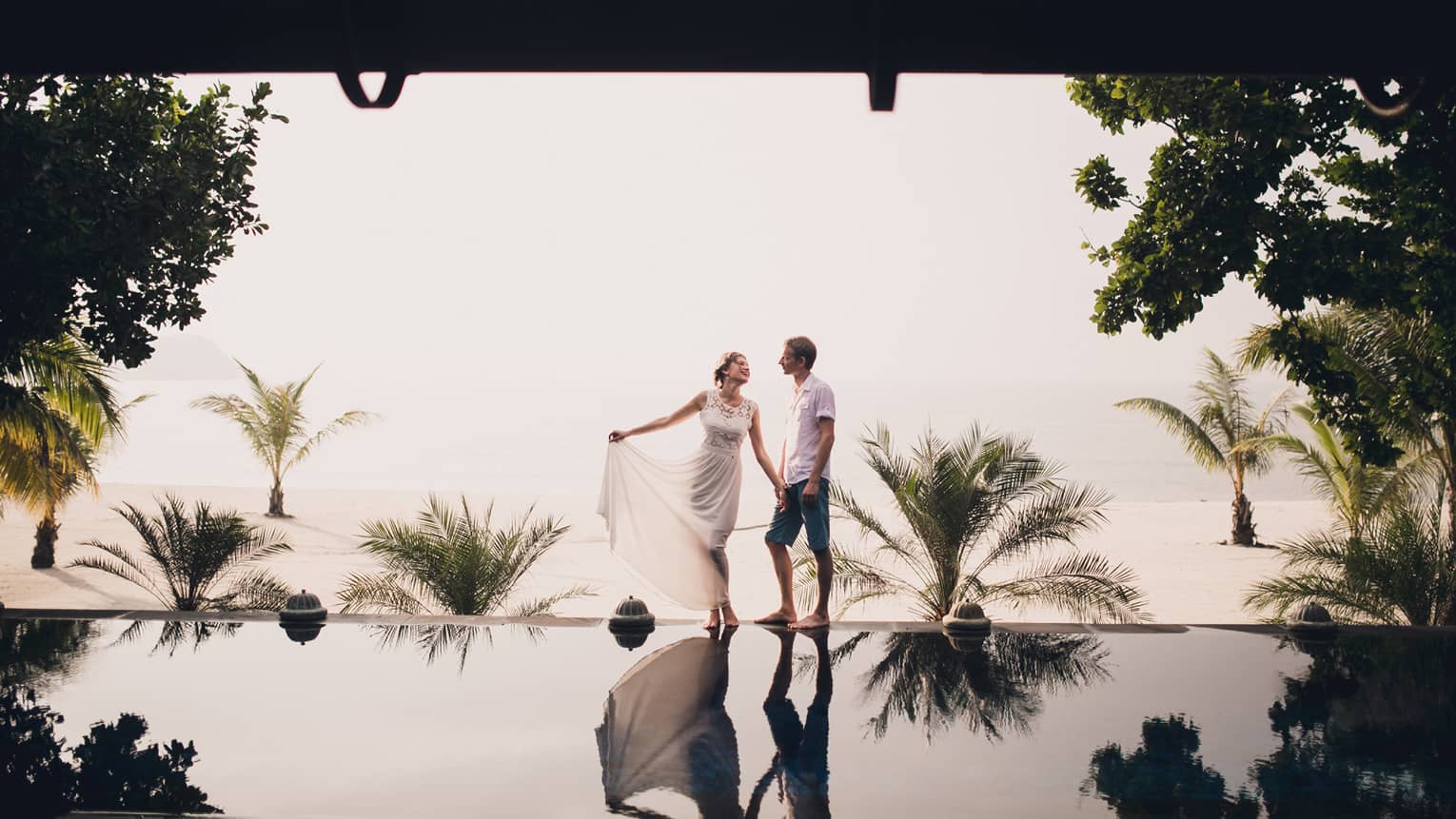 Woman in white lace dress and man walk along edge of reflection pool