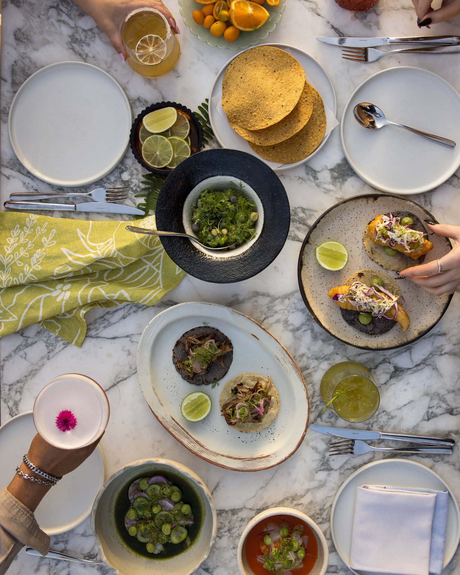 Overhead view of white marble table filled with plates of tacos, bowls of salsa, slices of oranges and limes, with two hands holding cocktails and another set of hands reaching for food