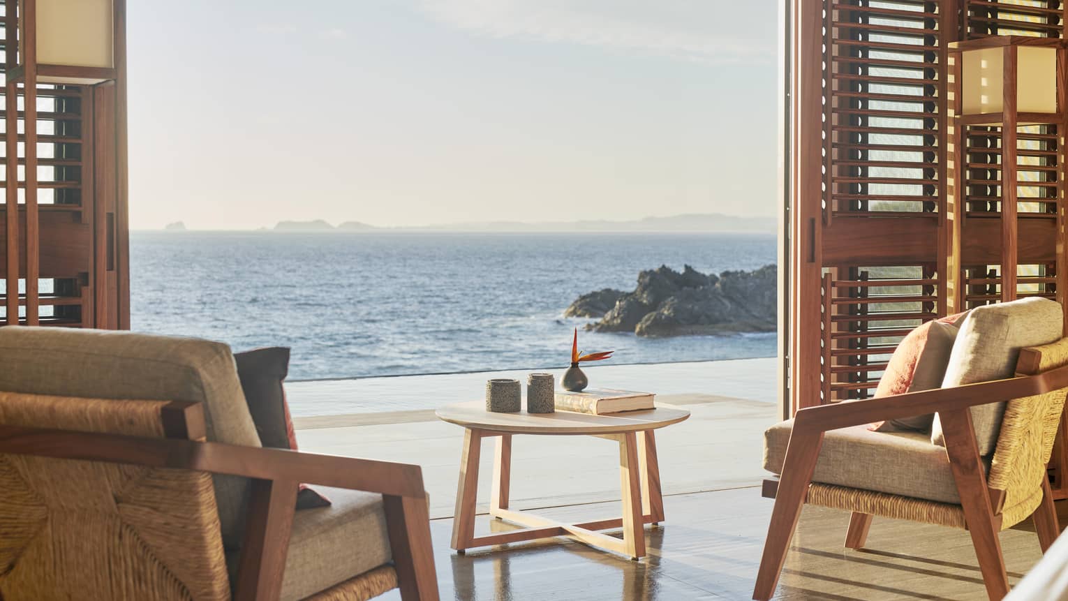 A serene seating area with wooden armchairs and a round table, overlooking the ocean. The open sliding doors offer a stunning view of the sea and rocky shoreline.