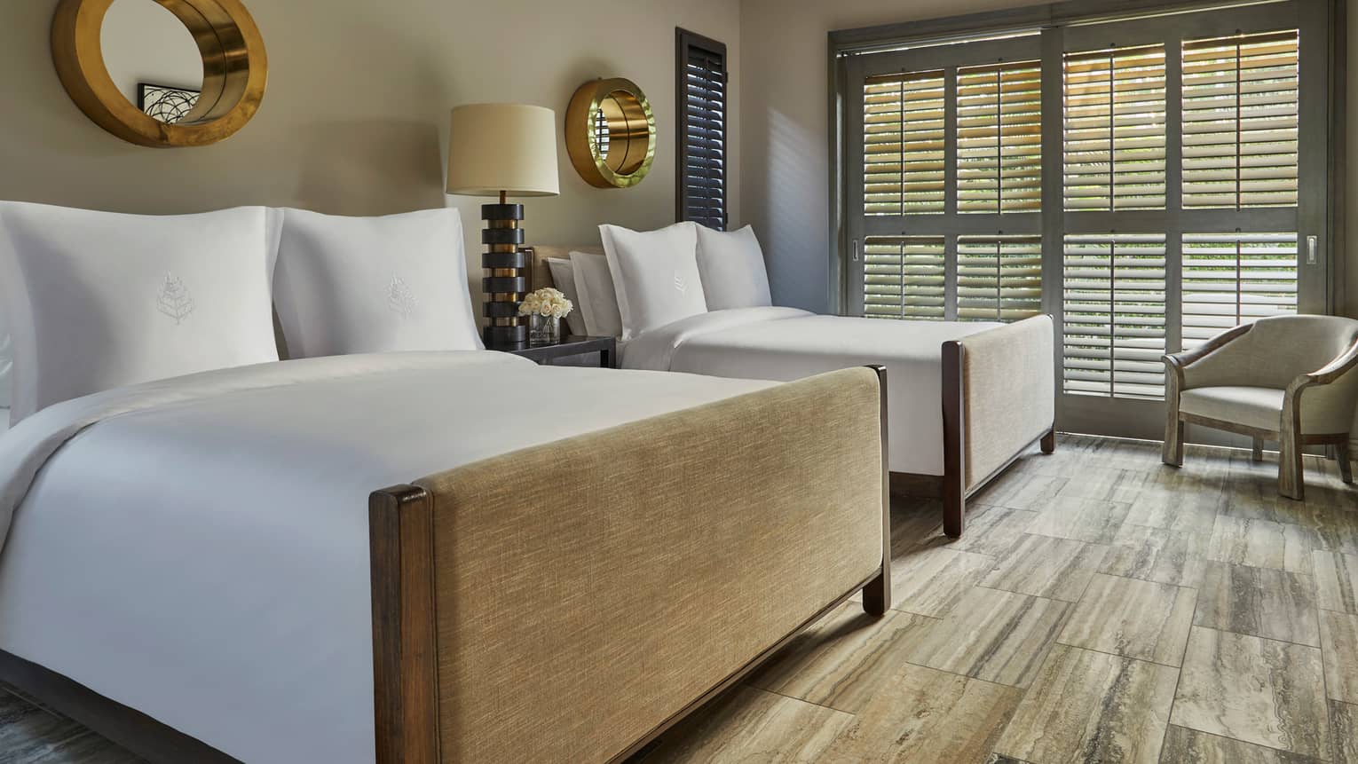 Luxury resort bedroom with two beds, white bedding, decorative mirrors, a nightstand with a lamp and shuttered windows letting in natural light