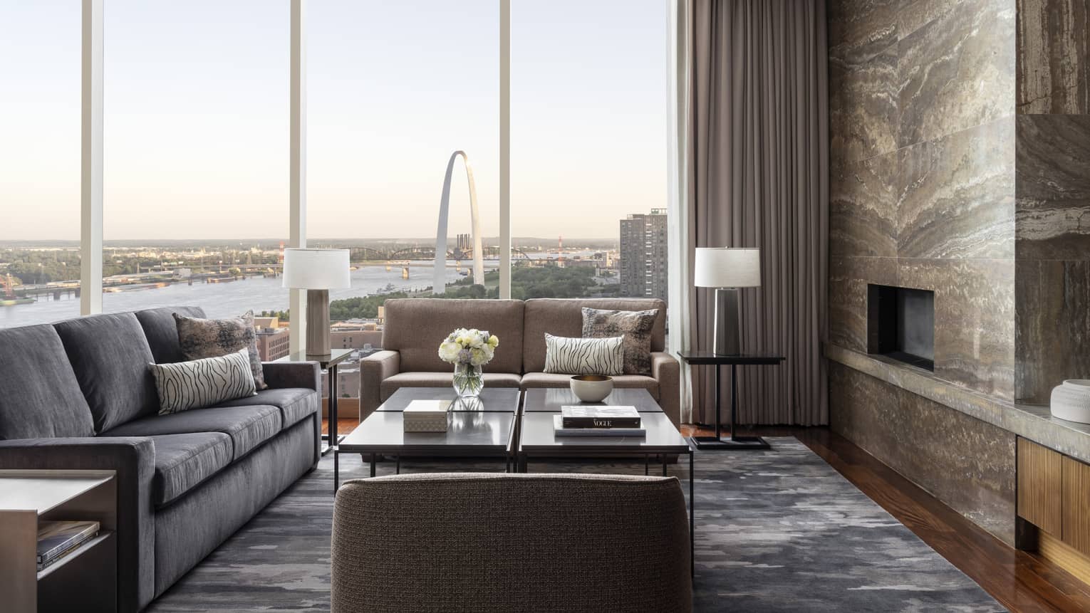 Living room with fireplace and stone wall, grey sofa, large square table, wall of windows with view of St Louis arch