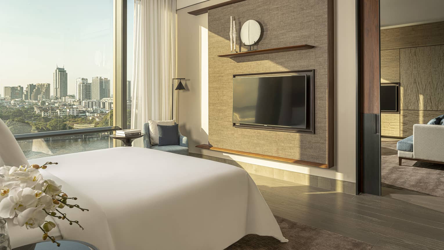 White king bed facing dark tan wall with TV, display shelf beside tall window with river and city view