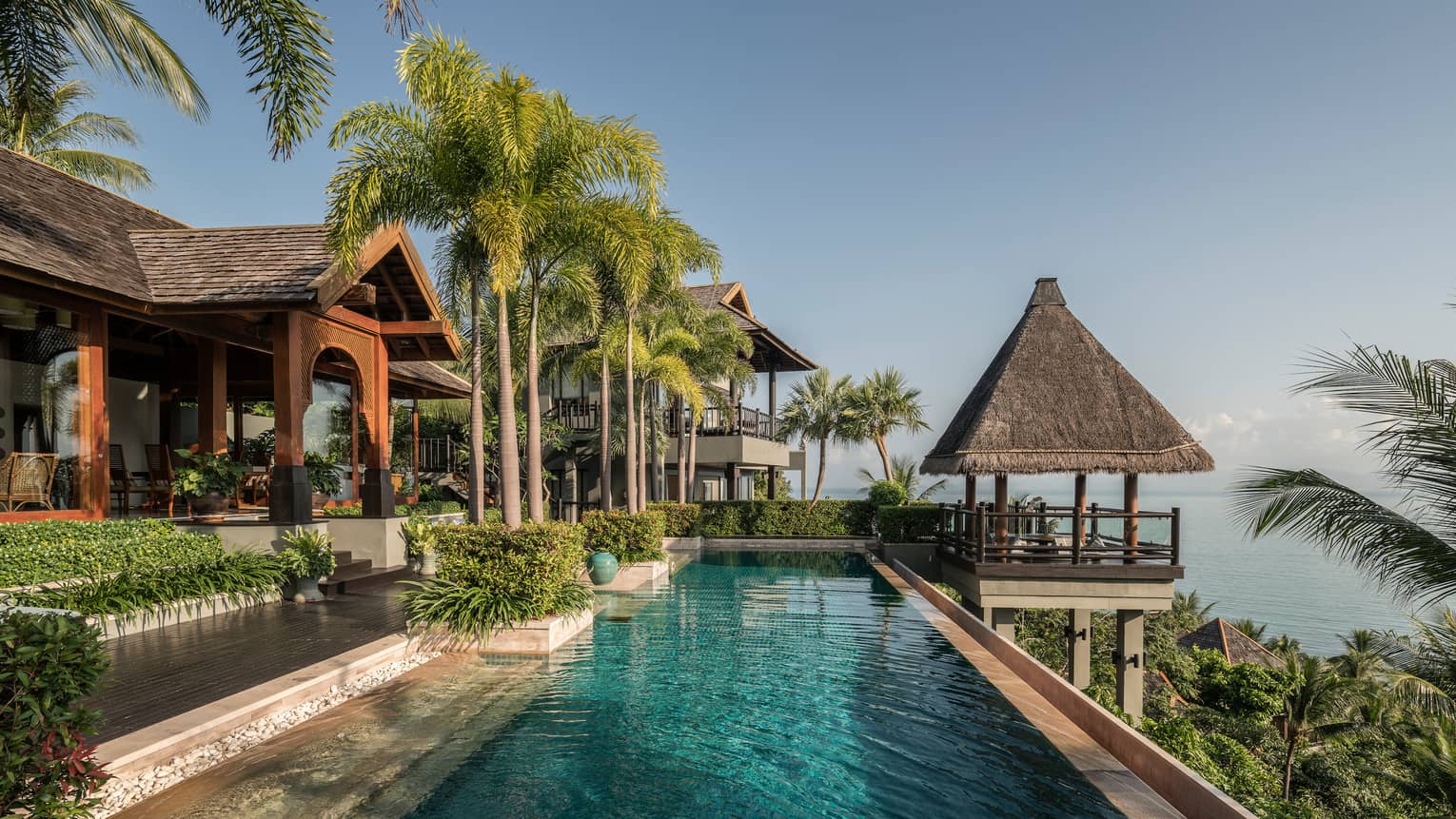 Long outdoor swimming pool under tall palm trees, thatched roof gazebo