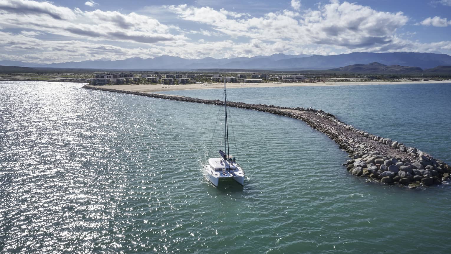 A sailboat sails toward the shoreline in the horizon and is surrounded by a stone walkway through the ocean