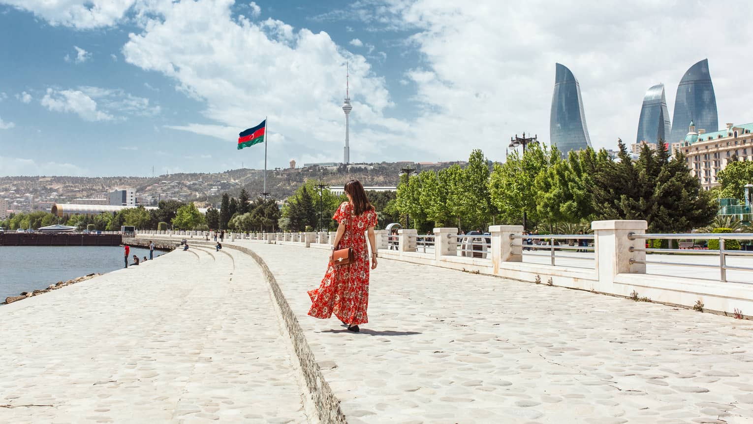 Woman wearing red dress walks along stone waterfront, skyscrapers in background