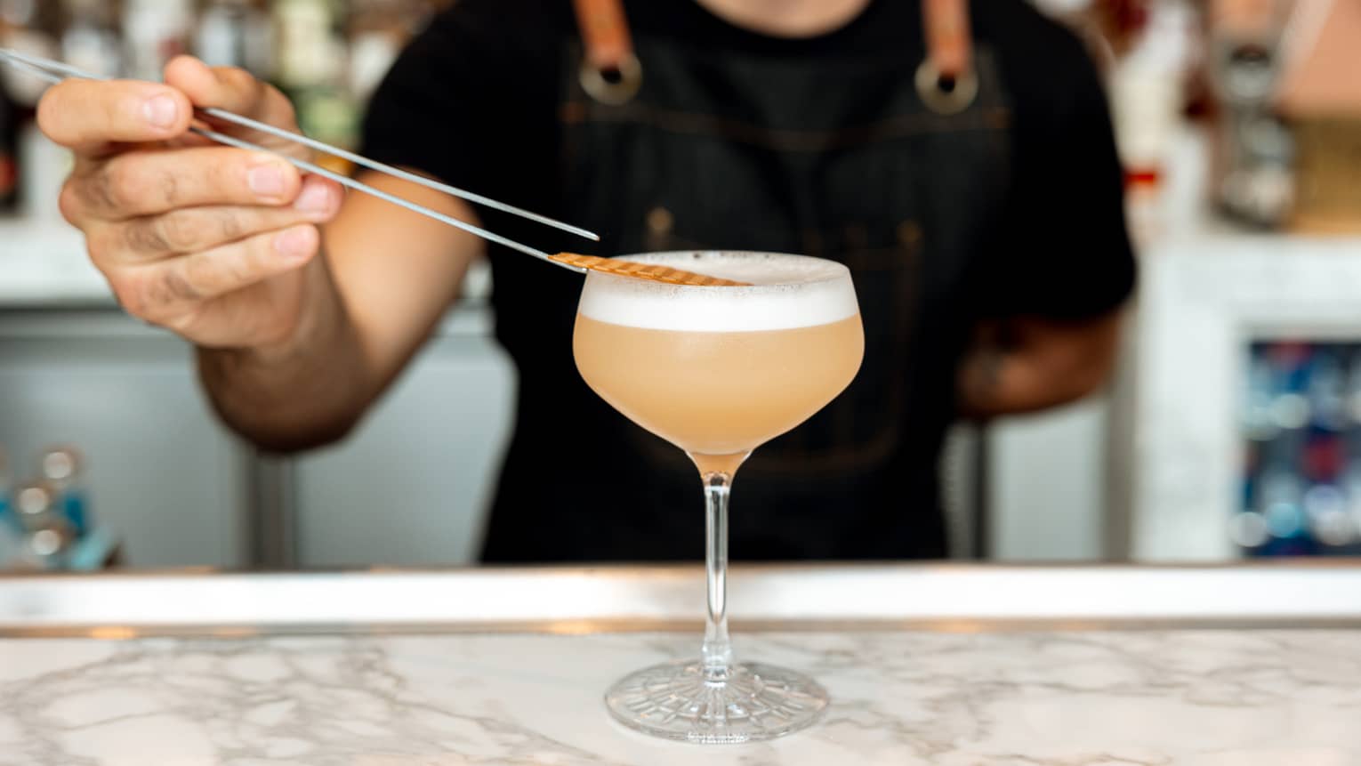 An apron-clad bartender uses tweezers to carefully garnish a frothy cocktail in a stemmed glass over a smooth, marble bar.