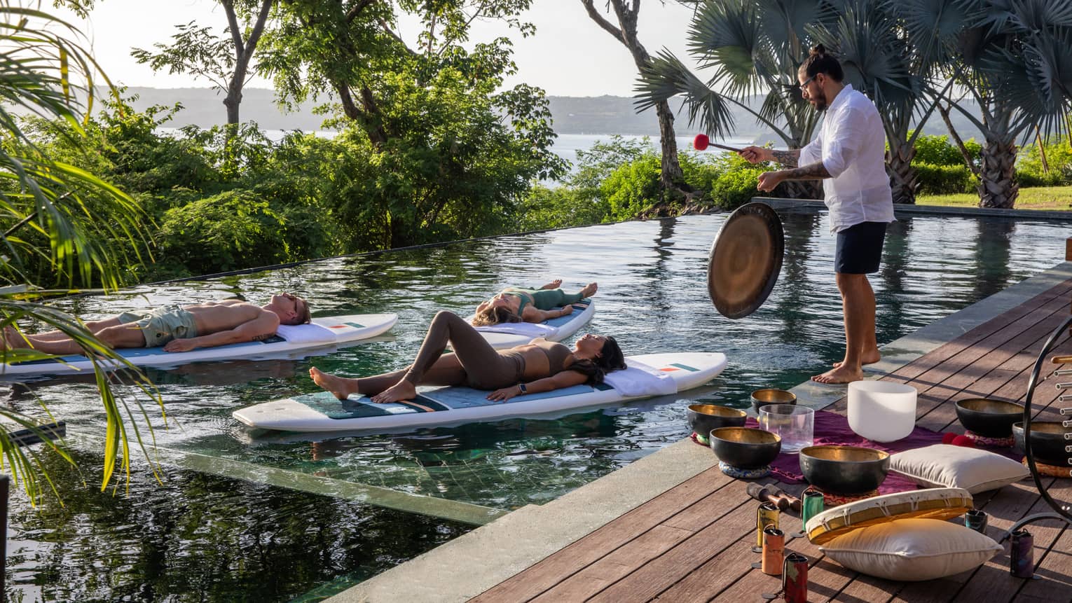 Three people float on stand-up paddleboards in a private pool while someone plays a gong on the deck