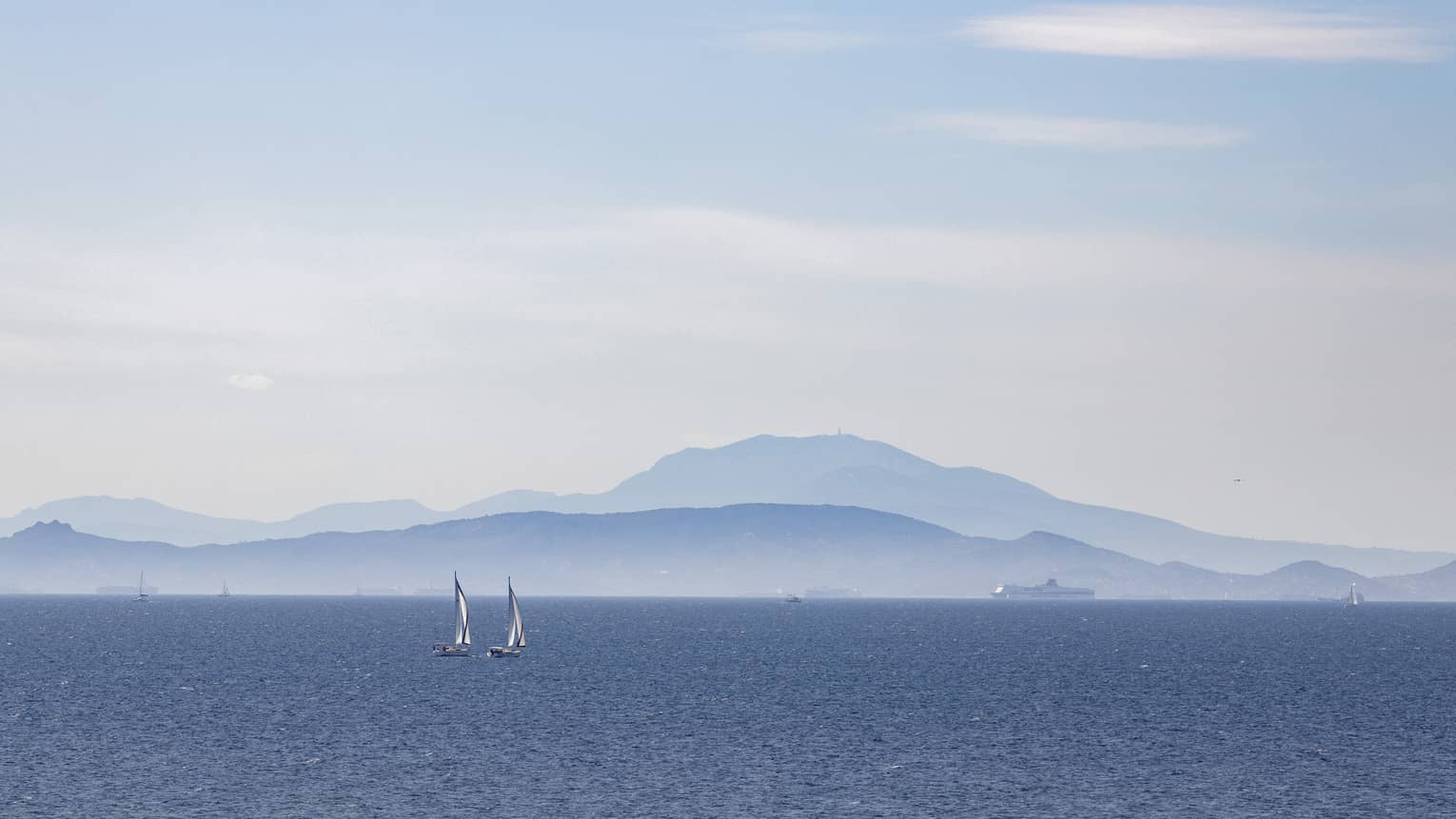 Two sailboats on blue ocean with blue-grey mountain range in the background 