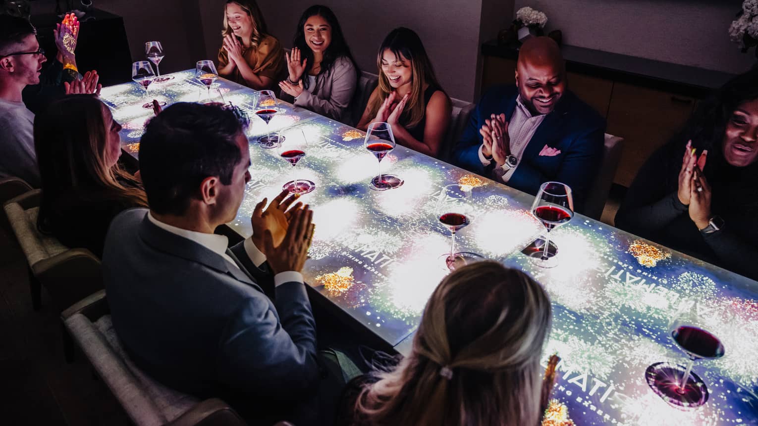 A large group of people sitting at a table with a screen on top of it drinking wine.