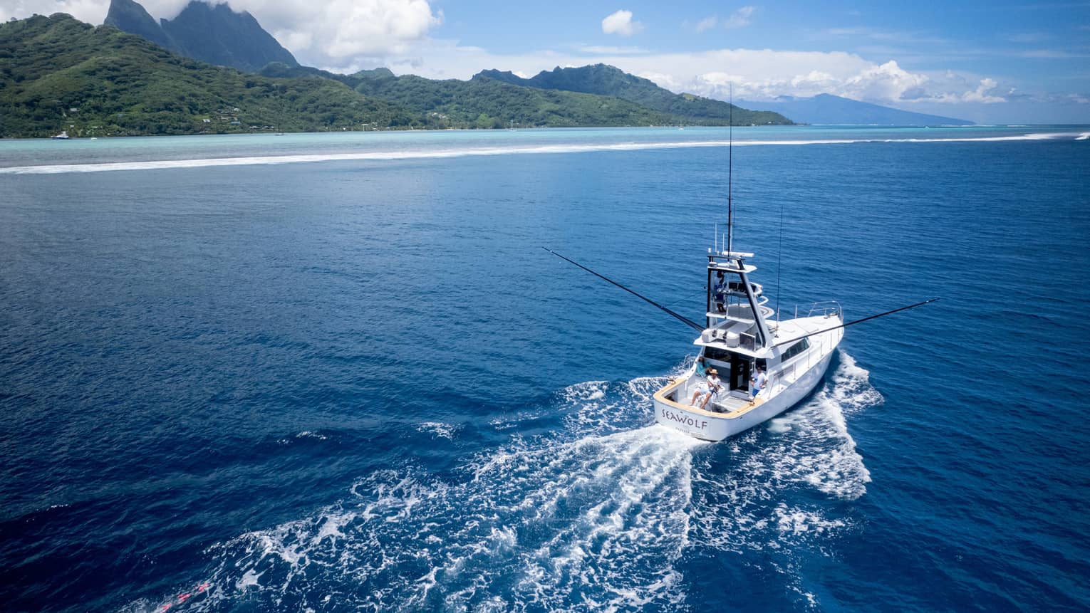 Yacht creates wake in water as it heads out to open sea, lush green mountains in background
