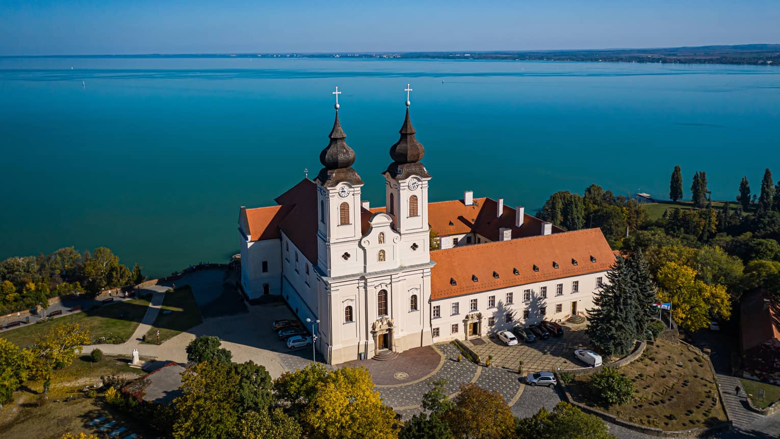 A Benedictine monastery, with white walls and a red tiled roof, sitting beside a large blue lake and surrounded by trees.