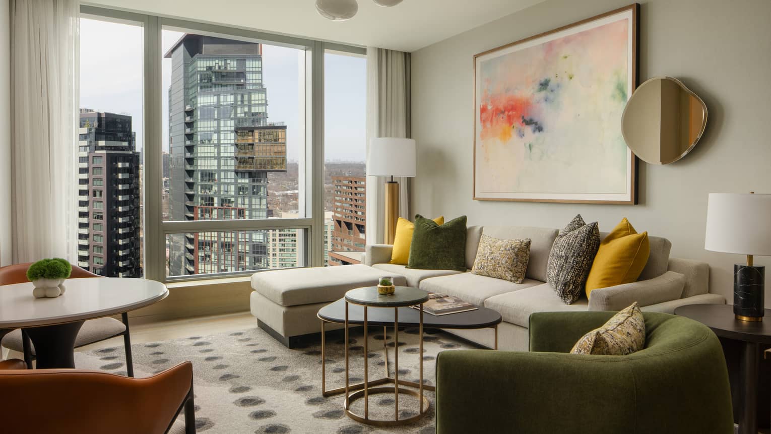 Stylish suite living area with a large sectional sofa, colorful pillows, modern artwork, and floor-to-ceiling windows overlooking the city