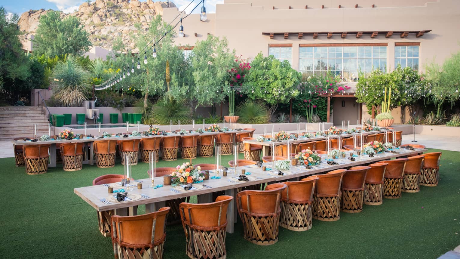 Three long tables outside with dark orange chairs on green grass next to a beige building.