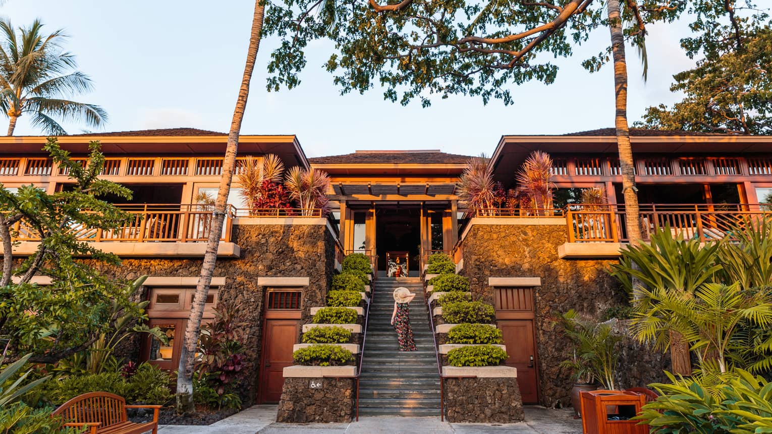Woman in sunhat walking up resort stairs with shrubs, two-storey building with balconies