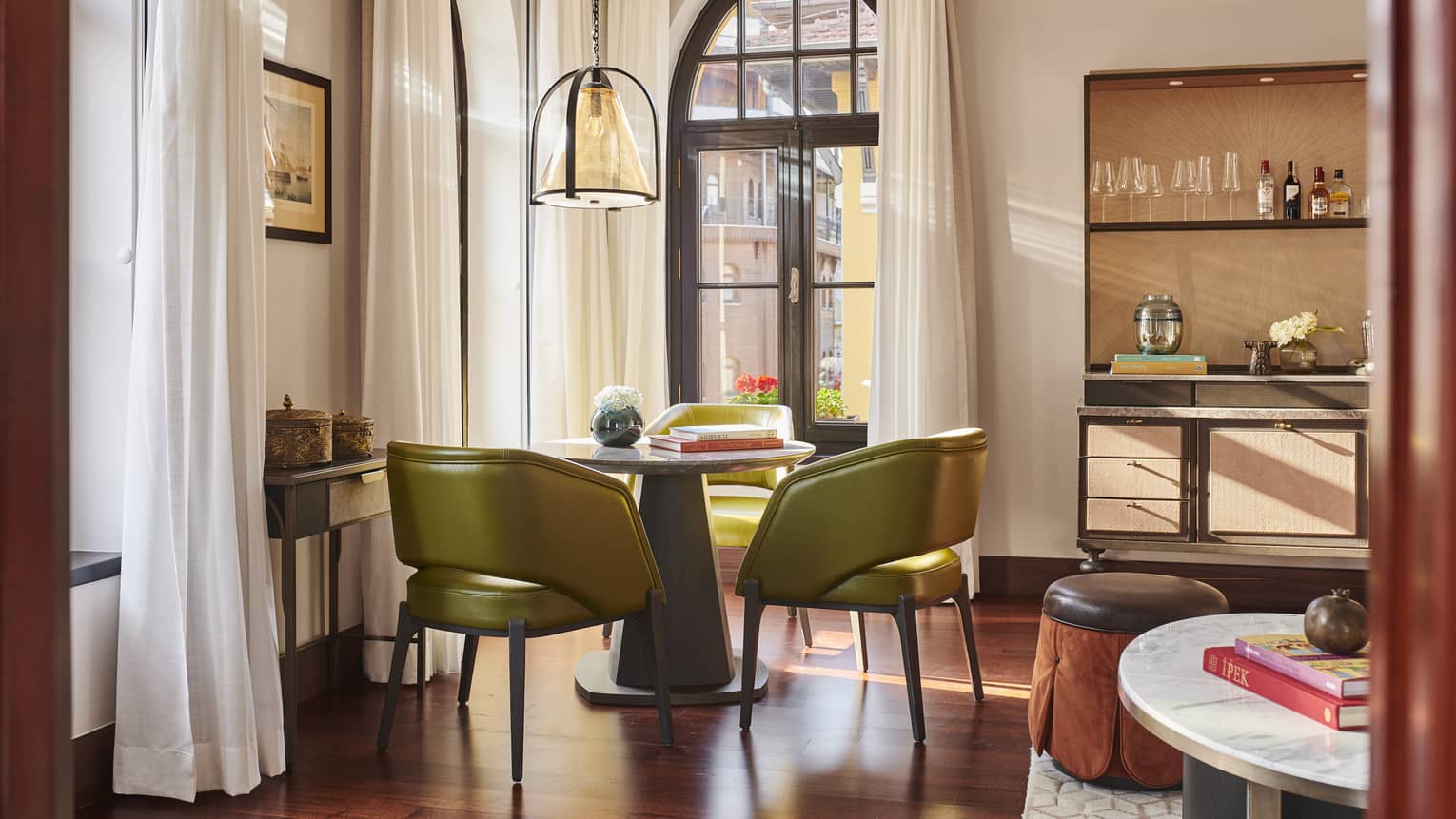 Hotel suite dining area with green contemporary chairs at round table and tall arched windows with long curtains