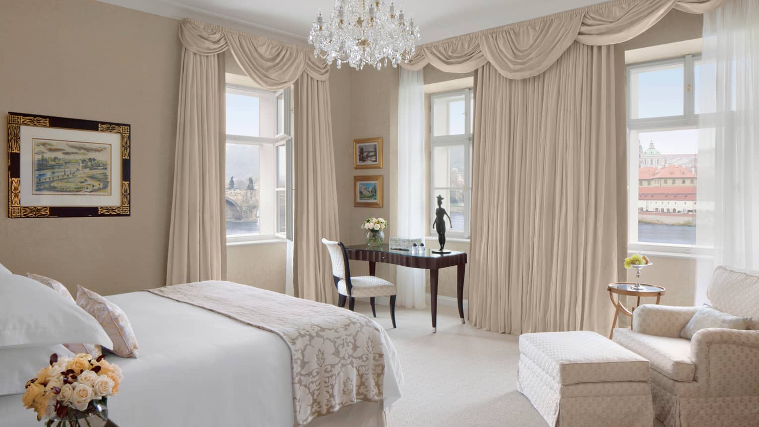 Room with white bed, tan club chair and ottoman, chandelier, tan curtains and desk
