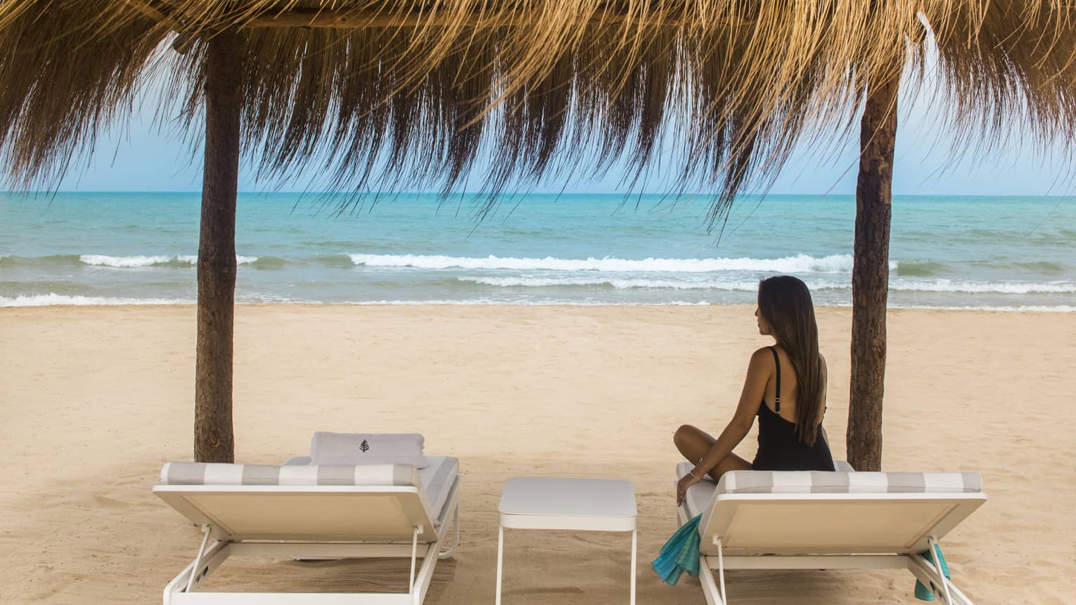 Long-haired woman in black swimsuit sits on lounge chair under grass-roofed beach cabana at Four Seasons Hotel Tunis