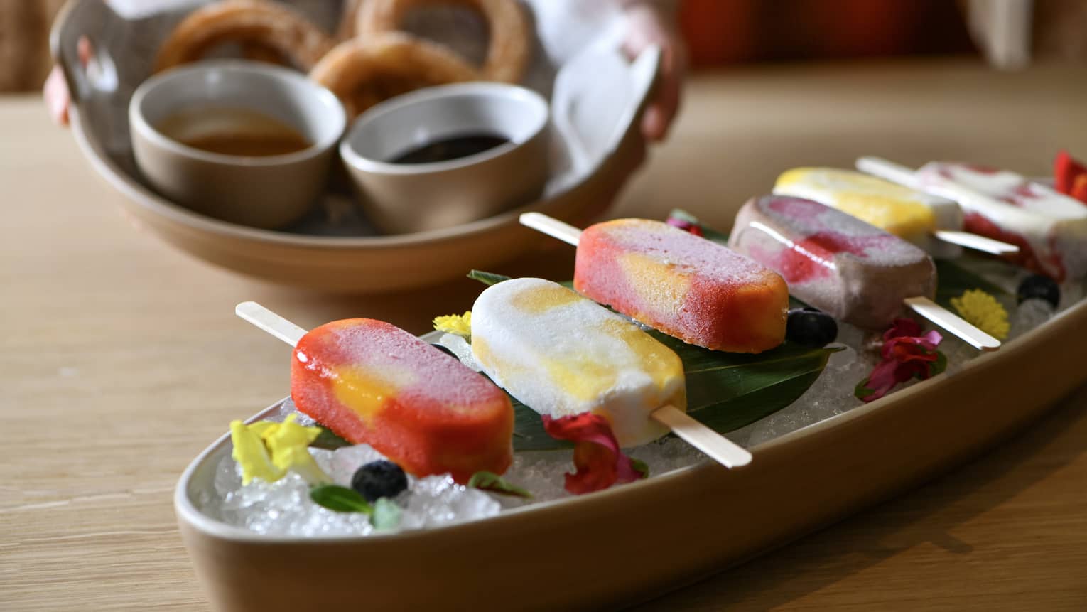 Six popsicles in alternating colors of red and white are lined up on a platter of ice and garnished with green leaves and red and yellow flower petals