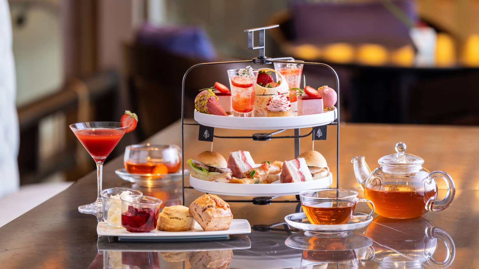 Two-tiered afternoon tea spread with sweets and savouries, glass pot of tea and cocktail in stemmed glass