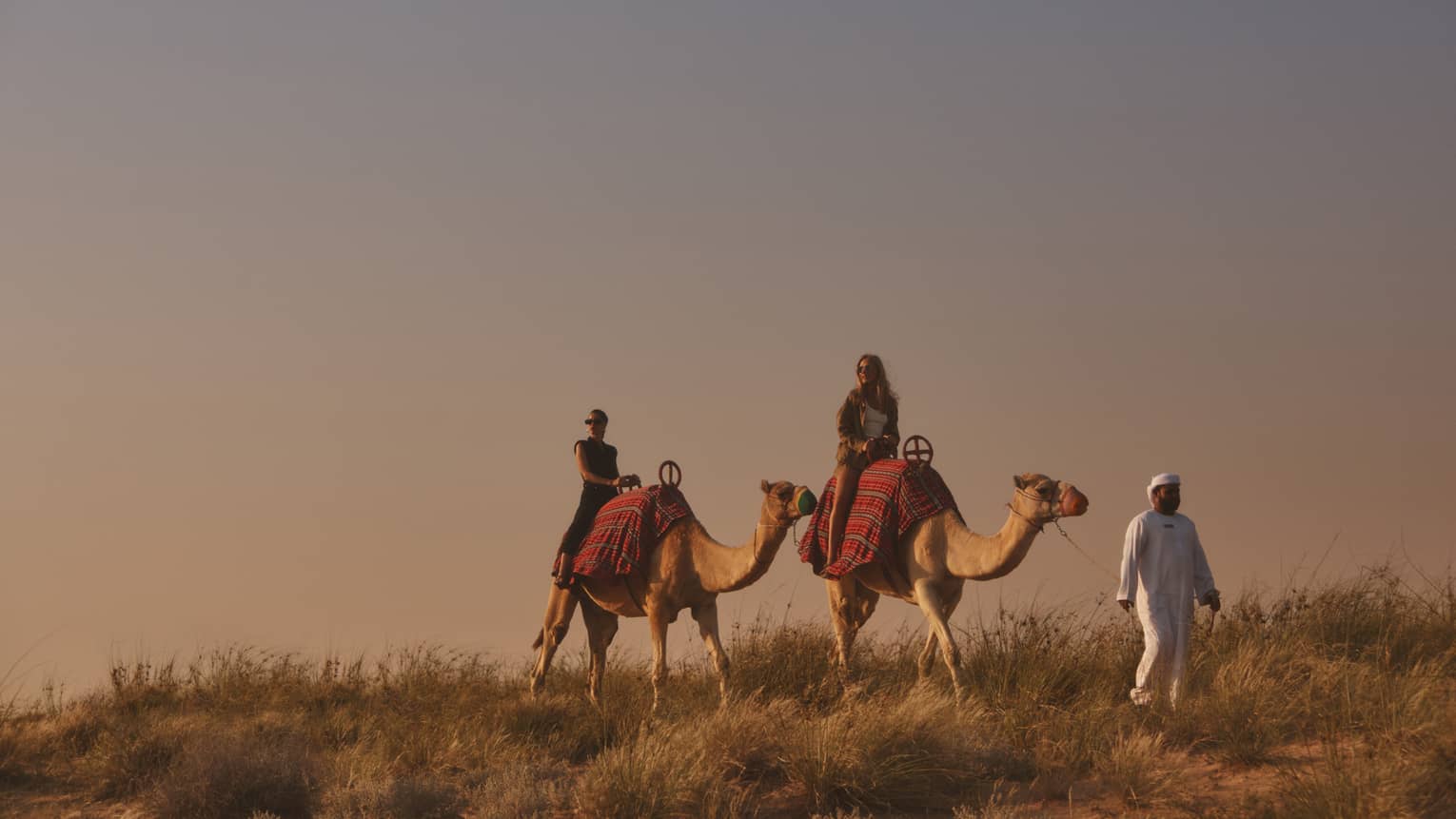 Two people riding camels with guide in front, leading them through a desert