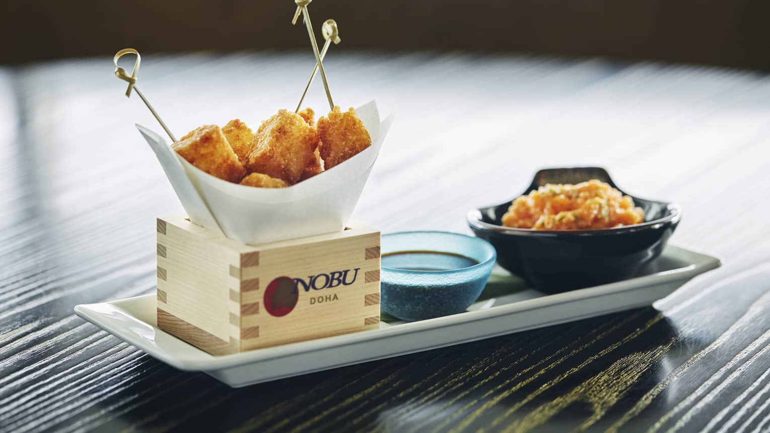 Close-up of white platter, wood box with Nobu logo, fried appetizers on toothpicks, dish with sauce