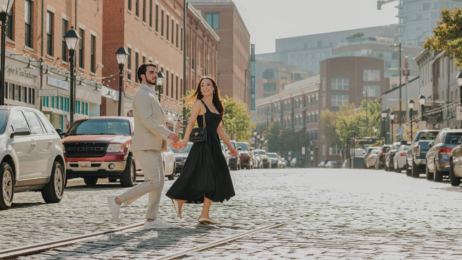 A smiling, well-dressed couple cross a cobbled street lined with stores in red-brick buildings, streetlights and parked cars.