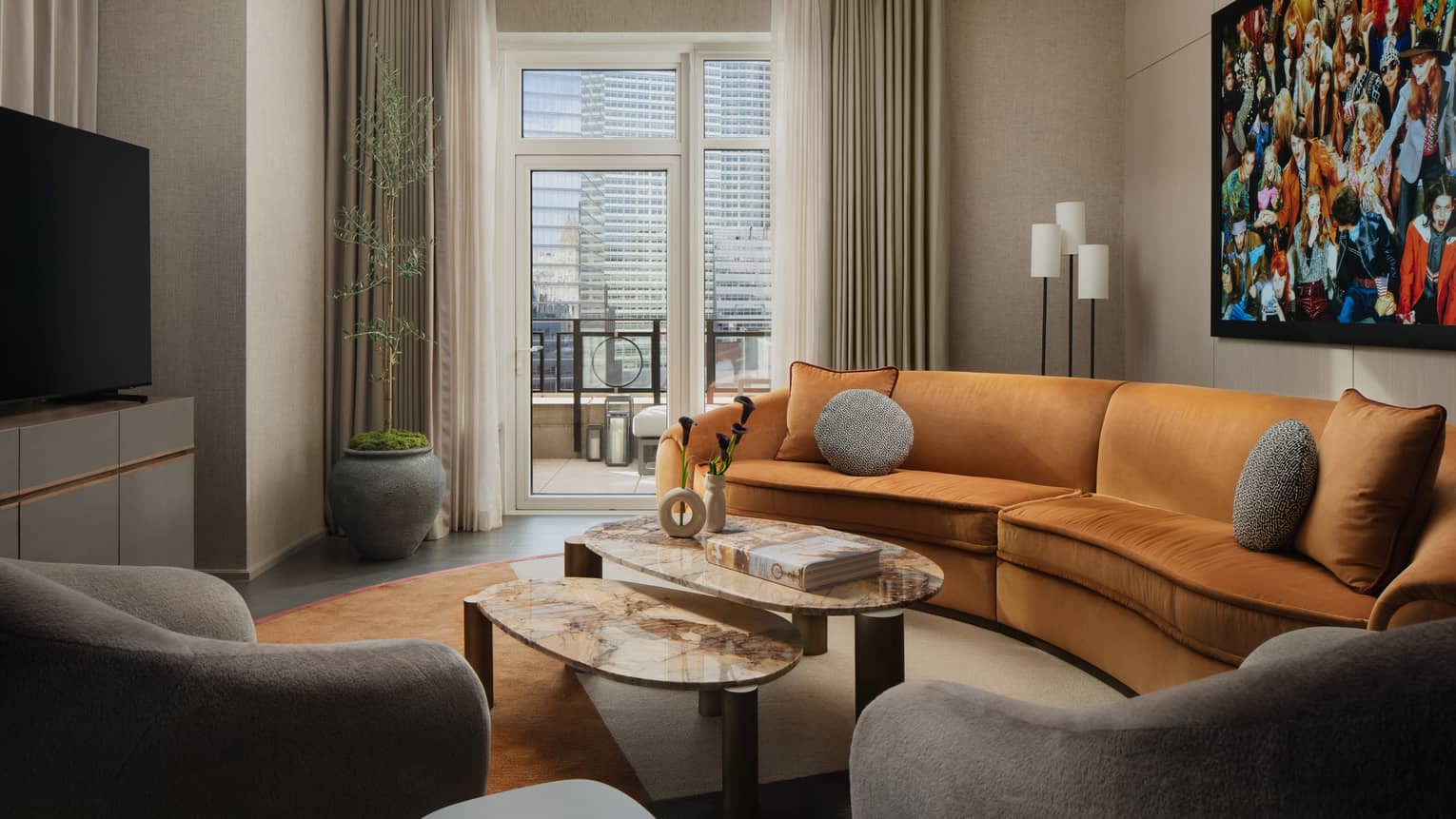 Hotel suite living area with curved orange sofa, coffee tables, gray armchairs, and colourful artwork on the wall, overlooking a balcony with city views