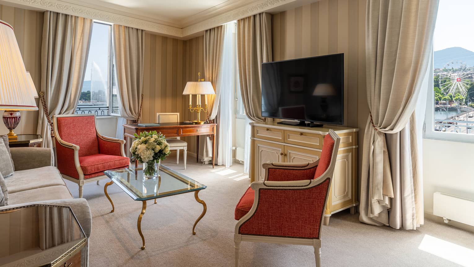 Elegant hotel suite with antique-style red armchairs, a glass coffee table, desk, sofa, TV, striped wallpaper and windows offering lake and Ferris wheel view