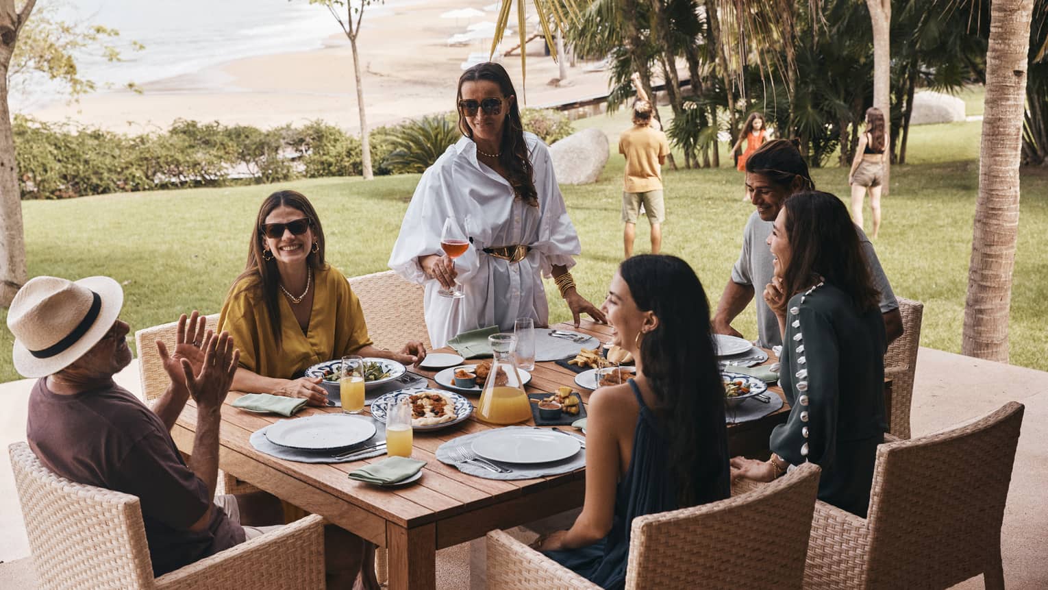 People seated around a square wood dining table in an outdoor area surrounded by trees and palm trees, with a lawn, beach and the shoreline in the background.