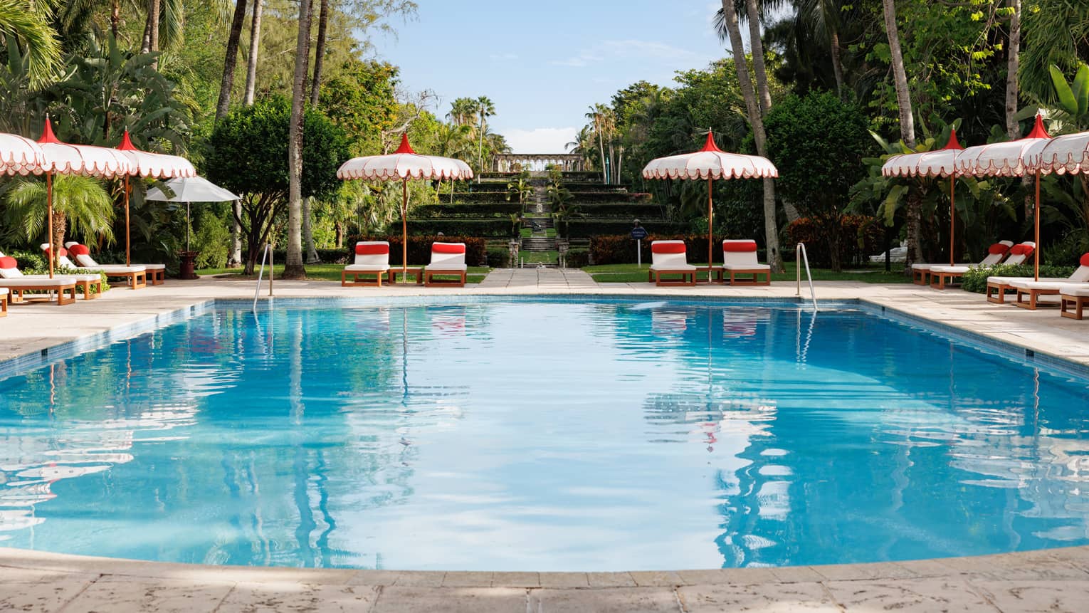 An outdoor pool at a tropical resort, flanked by white and red lounge chairs and umbrellas