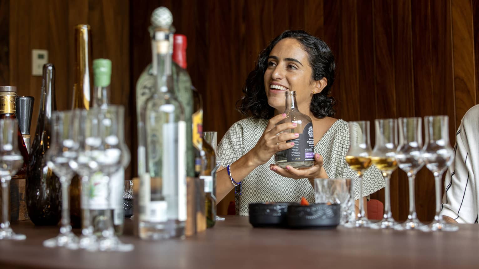 A guest cradles a short, wide bottle of liquor at a table littered with bottles of various sizes and grappa glasses. 