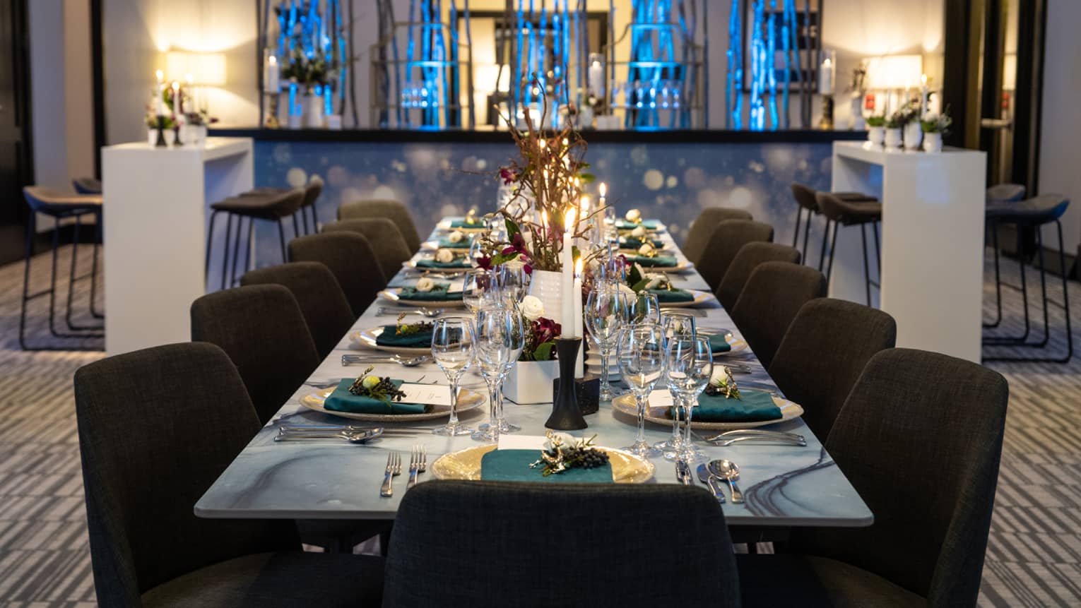 A long grey dining table with black chairs, teal napkins, and white plates; a bar is behind the table.