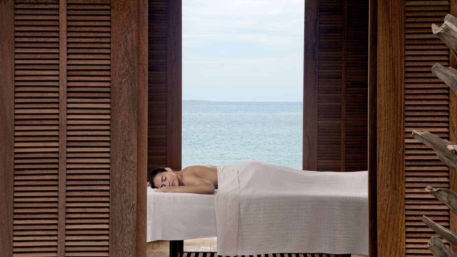 Guest enjoying a relaxing outdoor massage with a serene ocean view, framed by wooden shutters at a luxury beachfront resort