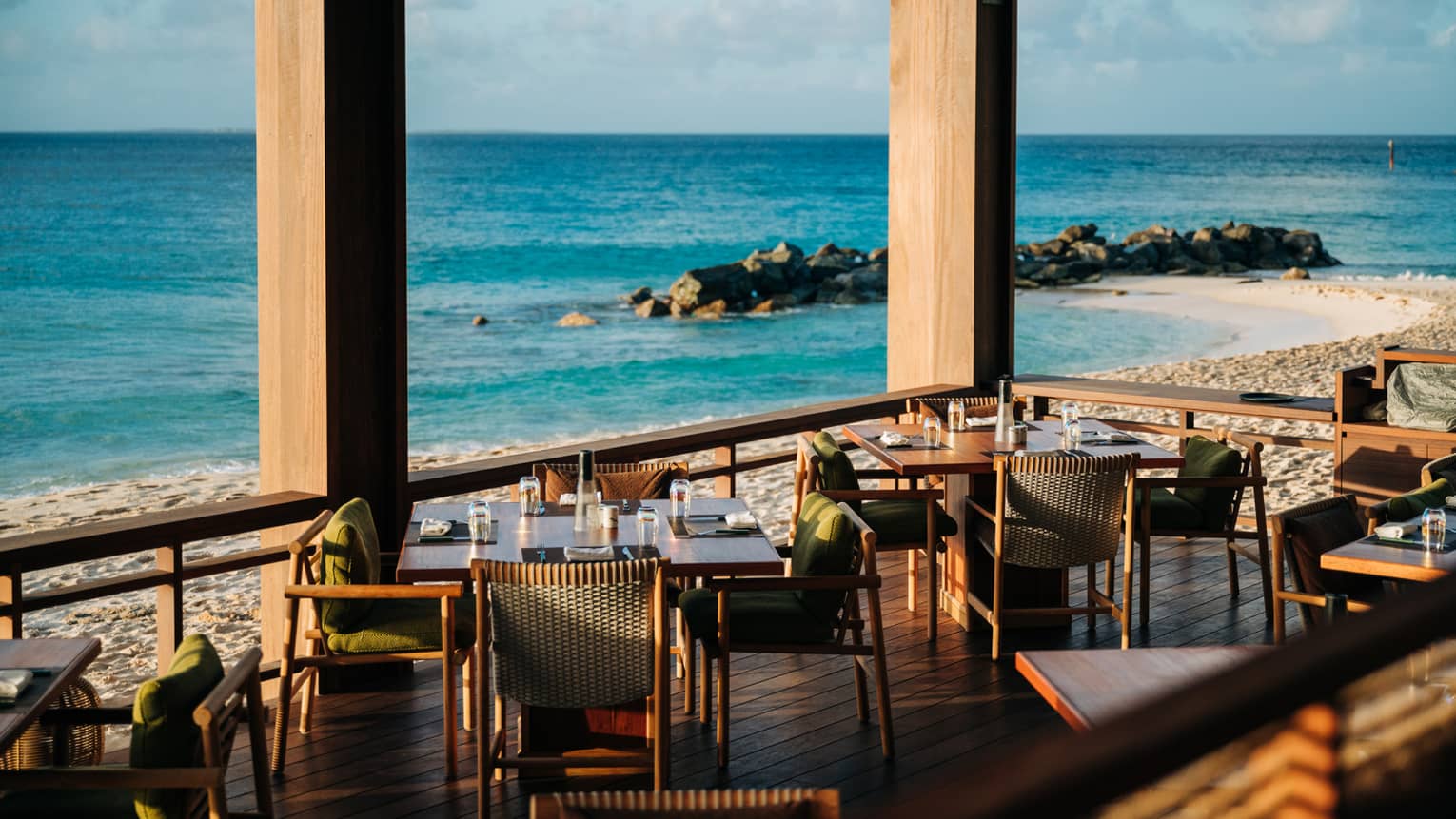 A covered but open-air outdoor dining area right on the beach, with wood deck flooring, tables, chairs, and amazing ocean views