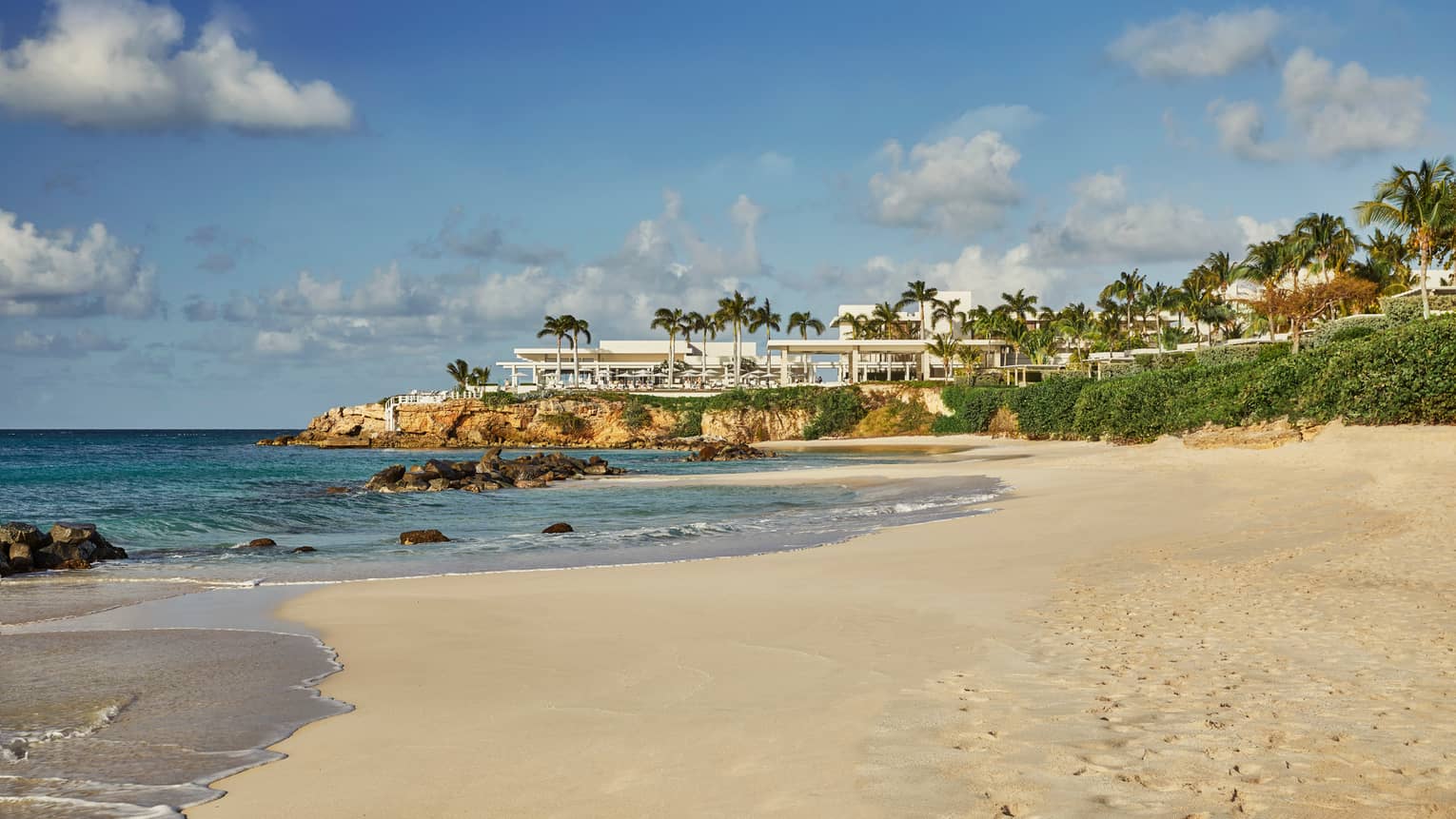 View across white sand beach to modern white event venue on rocks over sea 