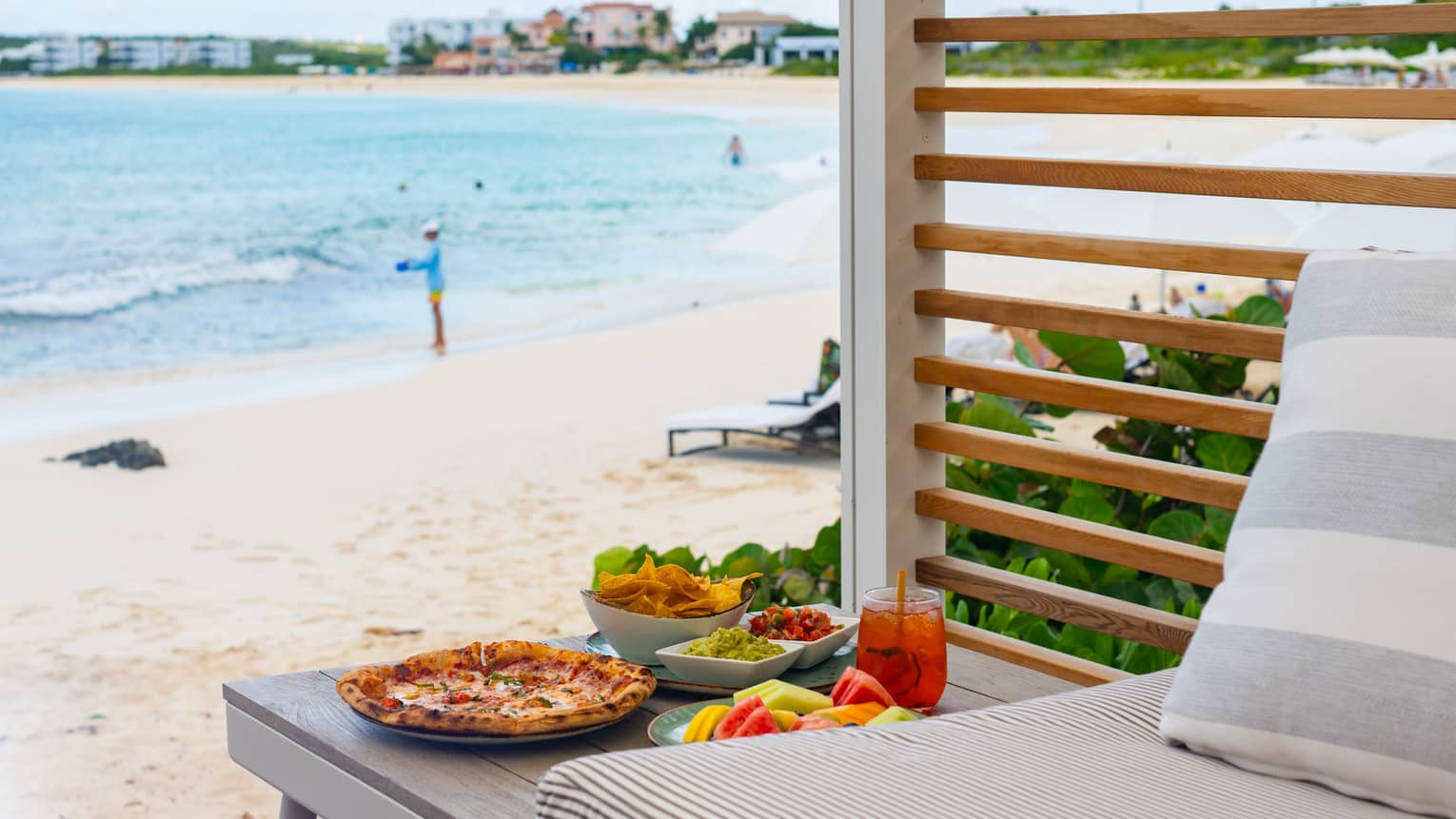 A table of pizza, cut fruit, chips and an iced beverage inside a beachfront cabana; in the background guests enjoy the ocean.