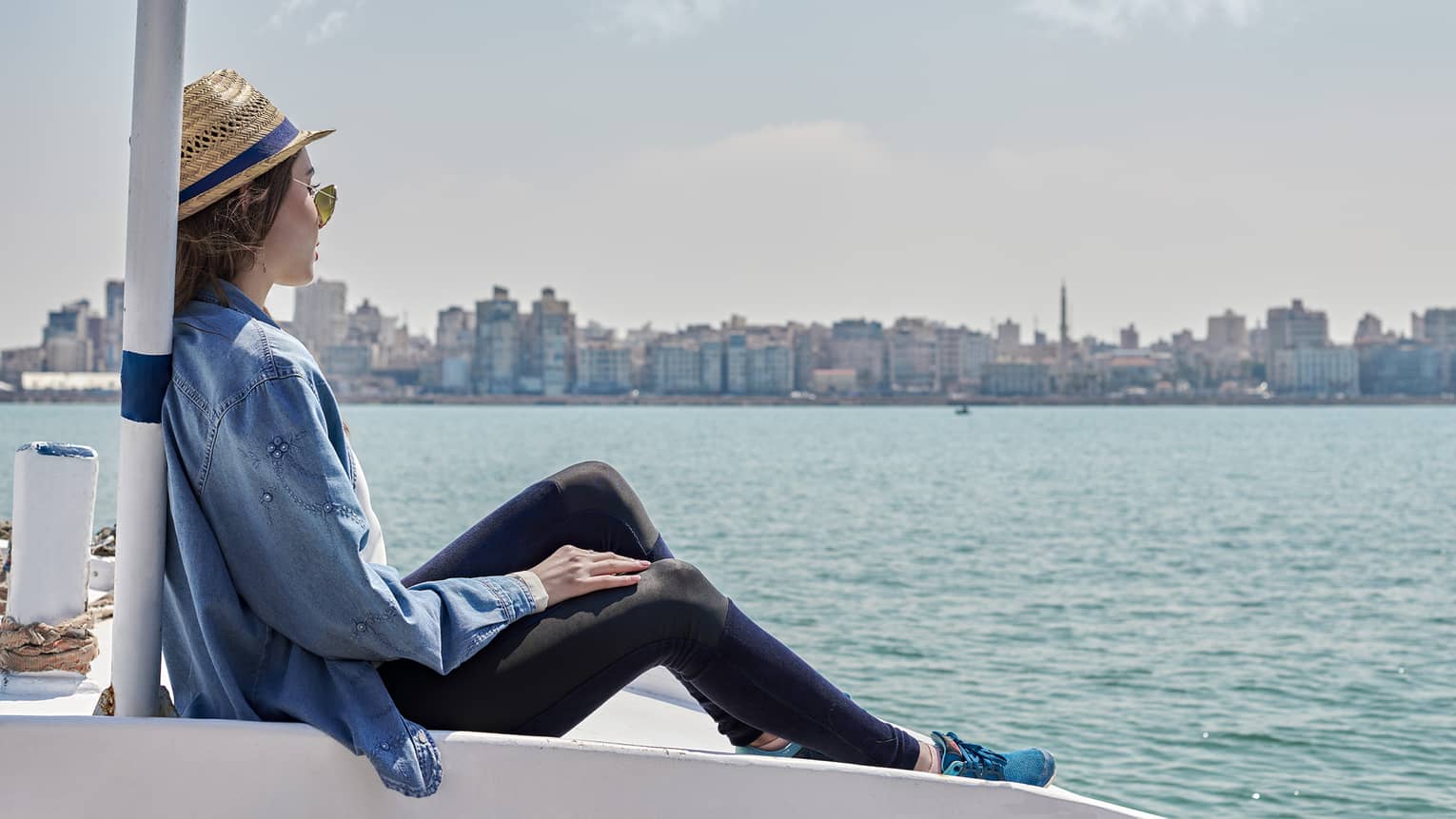 A guest wearing a straw hat sits on the bow of a boat and looks out over calm water toward a distant metropolitan shoreline.