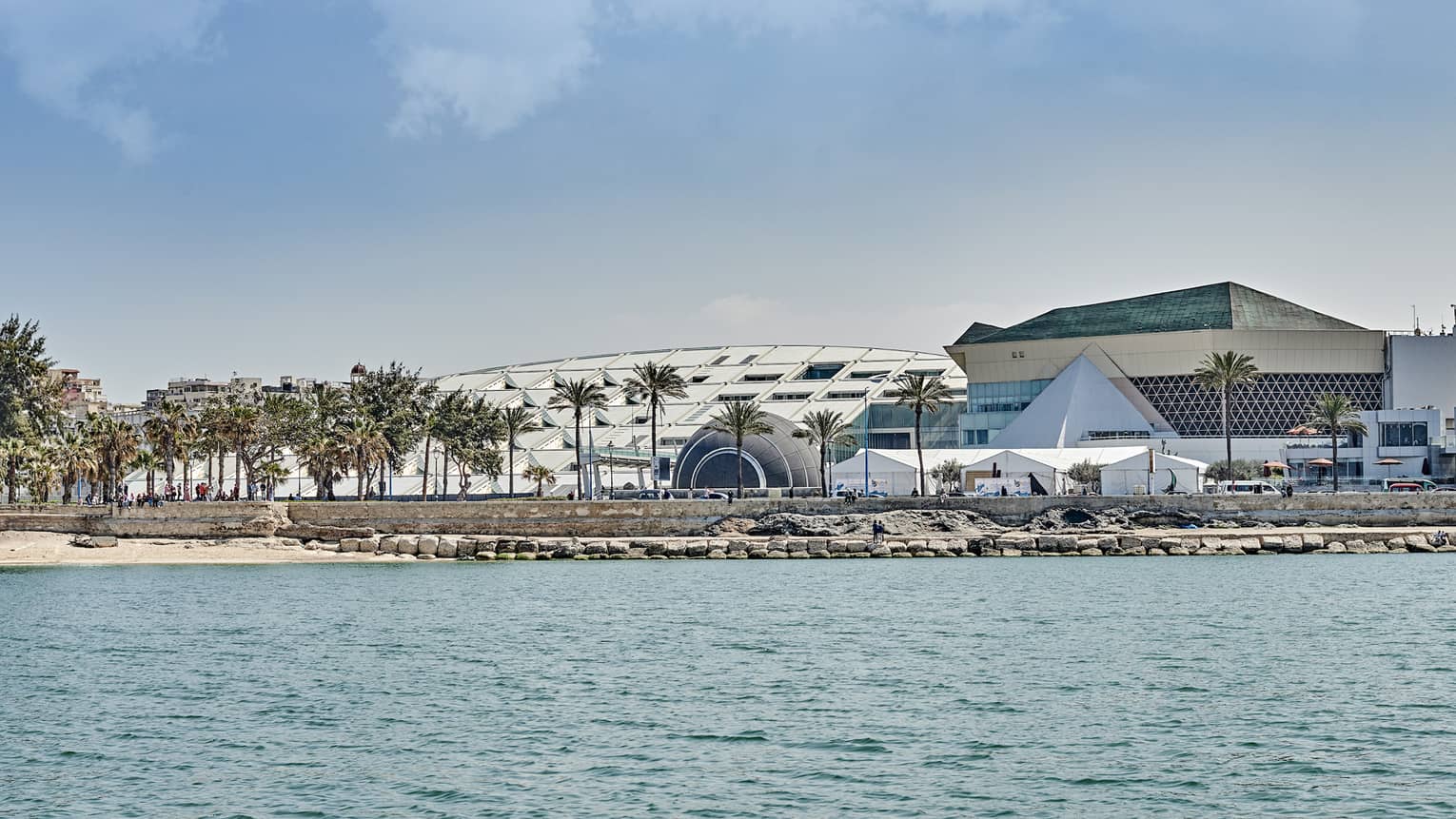View over calm water towards a stony shoreline where people mingle under palm trees in front of a large, sloped building.