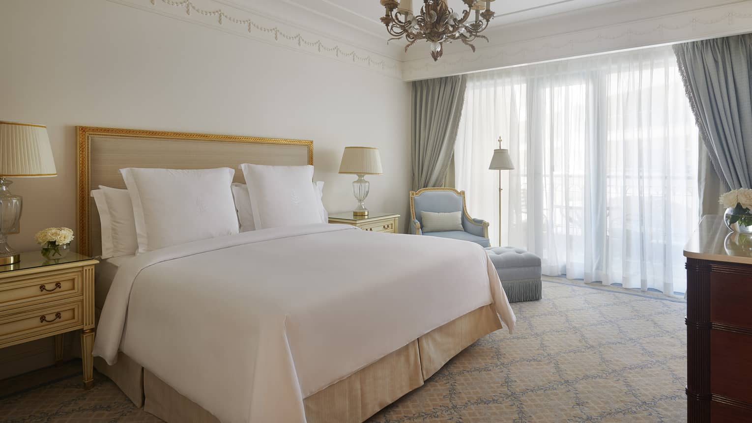 Suite with white bed, tan headboard, blue club chair and ottoman, chandelier, curtains